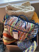 Box filled with various crocheted blankets and projects, including a cream-colored blanket on top and a rainbow striped blanket in a clear plastic bag.
