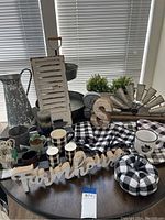 Wide shot of farmhouse decor items including metal pitcher, distressed shutter, windmill, wooden signs, candles, black and white checkered pumpkins, mugs, and faux greenery