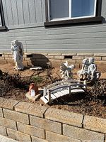 Photo of seven garden statues outdoors against a stone wall and dirt background includes a white angel, fairy, bridge, children on bench, with a terracotta frog on stone in front