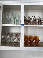 Cabinet shelf showing assorted glassware including clear etched glasses, green glasses, amber goblets, and four decorative beer steins with pewter lids.