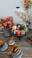 Glass bowl with stand filled with woven balls, small pumpkins, and flower stems. Three small orange flower baskets and grey planter with fall flowers.