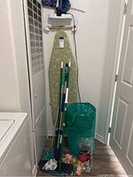 View of the ironing board mounted on the wall, multiple cleaning tools (mop, Swiffer), and a green laundry hamper on the floor in a laundry area.