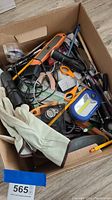 Box containing assorted junk drawer items including tools, office supplies, gloves, and small gadgets as visible from above.
