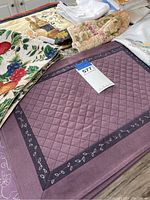 Photo showing stacked linens including white napkins, beige floral oven mitts, and quilted purple and fruit patterned placemats