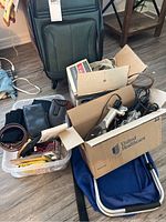 Small rolling black luggage suitcase, two cardboard boxes with assorted contents including hair styling tools and accessories, plastic container with belts and small bags, and a folded blue chair or bag on wooden floor.