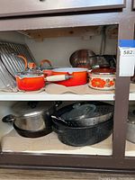 Shelf with orange Le Creuset pots and pans, ceramic floral pot, and stainless steel cookware underneath.