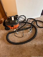 Black and orange RIDGID portable wet/dry vacuum with attached hose and power cord laid on carpet. View from side and front showing unit wheels and hose connection.