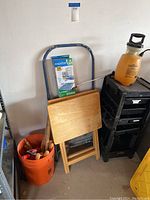 Photo showing wooden TV trays, a blue folding stepladder, orange bucket with wooden handles, black beauty container on wheels, yellow garden sprayer on top of beauty container, and boxed Magic Bags leaning against wall.