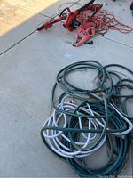 Electric blower, electric trimmer, hedge trimmer, orange extension cords, and garden hoses seen outdoors on concrete surface.