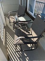 Two brown metal framed patio glider chairs with taupe mesh fabric seats and backs with glass top table between them, outdoor setting on balcony or porch. Small statue of a boy and dog atop table.