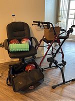 Photo shows black rolling chair with three bowling balls placed on its seat plus a foam roller. Black bowling ball travel bag on the floor contains another bowling ball. Red walker next to chair with cardboard box on its seat.