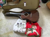 Washburn acoustic guitar in hard case, two holiday sweaters in front of case, on carpeted floor
