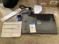 Overview showing HP laptop sealed in plastic, HP wireless mouse, power adapter, documentation and receipt on carpet floor