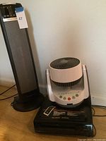 Three items grouped: tall black ceramic heater, white Hunter fan, and black Quasar VCR underneath.