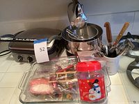 Photo of lot showing Cuisinart griddle, Sunbeam mixer with mixing bowl, bird toys in packaging, and cup with flatware including knives and forks.