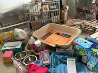 Photo of two large beige plastic dog litter boxes, pet bowls, cleaning products including Shock Clean, Insta Clean, and Scrubbing Bubbles, sponges and dust cloths, birdcage cover and pet exercise pen in background.
