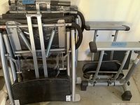 Photo showing two folded Malibu Pilates exercise machines with metal frames and black padded areas stacked and leaning against a wall.