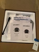 Opened box showing Kitchen HQ Air Grill securely packed in white styrofoam with power cord, metal skewers, instruction manual, and recipe book on top.