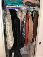 Photo showing assorted ladies coats and jackets hung on rack inside a closet, including faux fur coats and padded jackets.
