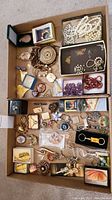 Top view of two boxes containing mixed costume jewellery including pearl strands, beaded necklaces, brooches, pins, keychains, and chains in different styles and colors.