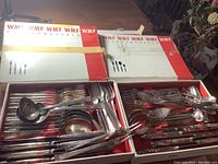 Two open boxes show a variety of forks, knives, spoons, ladles, and serving utensils arranged inside amidst red trim lining.