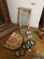 Photo showing small metal frames with vintage-style painted artwork, two porcelain ashtrays with blue and white floral designs, a clear square glass box with lid, Pearl brand glass washboard standing upright, and fabric table mat partially visible.