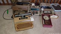 Photo showing a beige Coronamatic electric typewriter and vintage black Underwood manual typewriter side by side, other items including adding machine, clocks, and bathroom scale in the background.