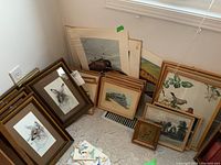 Corner area with a collection of framed art resting against wall including wildlife watercolours, railway sketches, and large lithographed print of birds in wood frames.