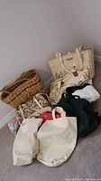 Grouped assortment of eight tote bags on floor against a wall. Includes natural woven rattan, green woven, several canvas and cotton bags in cream, white, black, and colorful prints.