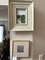 Two framed artworks hanging on a wall vertically. Top is a small, rustic framed oil painting landscape. Bottom is a larger framed square floral artwork.