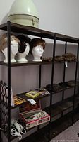 Four foam wig stands with eight wigs and hair pieces displayed on black metal and wood shelving unit, along with hair cutting kit and various small hair accessories spread on lower shelves.