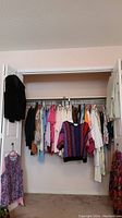 Wide view of a clothing rack holding a variety of women's tops, sweaters, blouses, and a few dresses. Clothes are mixed colors and patterns, hanging on hangers. Some extra tops/dresses hung on door and wall.