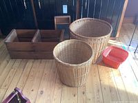 Photo shows two large wicker baskets, wooden washboard, wood packing crate and small red resin basket on wooden floor.