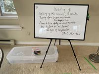 Whiteboard with handwritten quote positioned on a black tripod stand. Markers in plastic bag on clear storage container beneath the whiteboard.