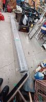 Long roll of grey carpet wrapped in plastic, situated in a workshop or garage with tools and equipment in the background.