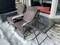 Two metal frame patio chairs with grey cushions and a matching patio table with square glass top. Items placed outside on a snow-covered deck.