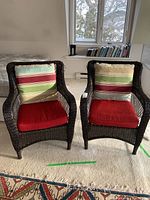 Two matching dark brown resin wicker chairs with cushions featuring a striped pattern on the back cushions and solid red seats placed on carpet.