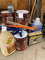 Overview photo showing the Wood Shield oil stain cans, carpernter glue jug, caulking tools, spray paint kit, and windshield washer fluid, placed on concrete floor in a garage environment.
