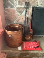 Photo showing the full fireplace tool set including the copper bin, log tongs, fire blanket, wooden-handled broom, and metal dipper arranged beside a stone hearth.