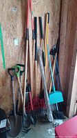 Photo showing an assortment of garden tools leaning against a wooden wall in a shed, including spades, rakes, hoes, and a turquoise dustpan and broom set