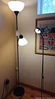Full view of the torchier floor lamp with goose neck side lamp next to a wall and power outlet, showing black metal pole and base with white bowl shade and smaller goose neck lamp attached.