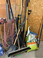 Photo of various outdoor tools stacked against a wall including shovels, rakes, brooms, and bags of lawn fertilizer.