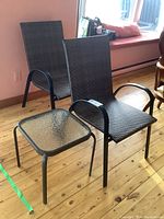 Two black resin wicker patio chairs with black metal frames and a small glass top side table on a wooden floor.