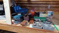Full lot photo showing the three sanders and various sanding belts, discs, and dust bags on a wooden shelf.