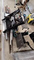 Top view of assorted scrap metal pieces including a large black metal frame and rods on garage floor with other items around.