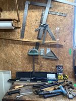 Partial view of wall and table showing various measuring tools, hatchet, crow bars, hammers, tape measure, and clamps.