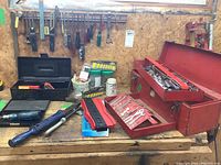 Wide view of workbench showing multiple screwdrivers hanging on the wall, red metal toolbox open with sockets inside, and black plastic toolbox on the bench.