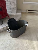 Oval metal tub with black metal lid inside, positioned near a hearth on a beige carpet.