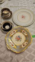 Photo showing two teacups with three saucers, one black and gold pattern, one different pattern. Dinner plate and serving platter visible.