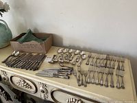 Full layout of flatware pieces on a dresser surface with a cardboard box to the left.